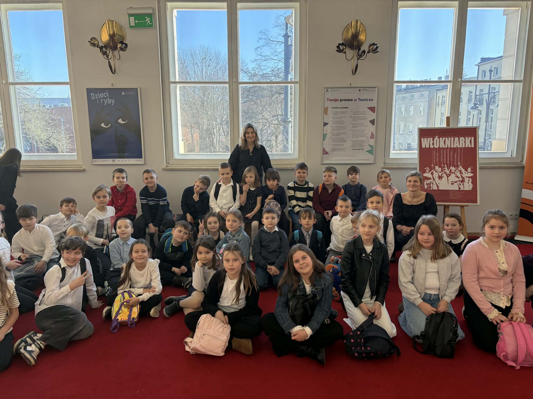Uczniowie klas 1b i 3b wraz z opiekunkami siedzą na czerwonej wykładzinie w holu Teatr Arlekin po obejrzeniu spektaklu „Dzieci i ryby”. Dzieci pozują do wspólnego zdjęcia przed dużymi oknami, przez które wpada jasne światło dzienne. W tle na ścianach widoczne są plakaty teatralne oraz dekoracyjne lampy, a cała grupa uśmiecha się do zdjęcia, tworząc pamiątkę z wizyty w teatrze.