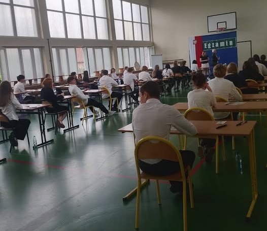 Uczniowie klas &oacute;smych podczas egzaminu z języka polskiego w szkolnej sali gimnastycznej. Na zdjęciu widoczni są uczniowie pracujący w skupieniu nad arkuszami egzaminacyjnymi.