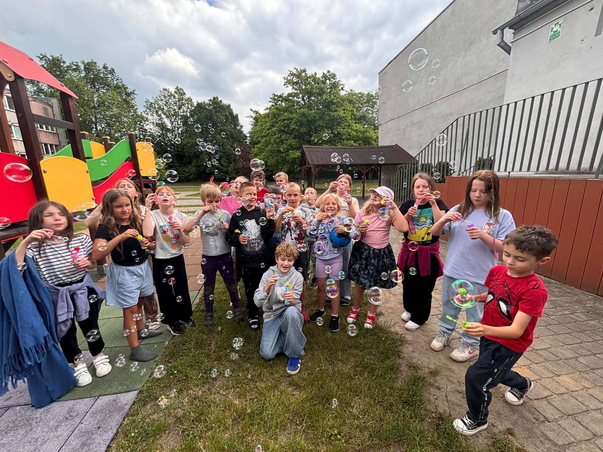 Grupa uczniów podczas zabawy z bańkami mydlanymi na szkolnym placu z okazji Dnia Dziecka. Dzieci stoją przed placem zabaw i tworzą kolorowe bańki unoszące się w powietrzu.
