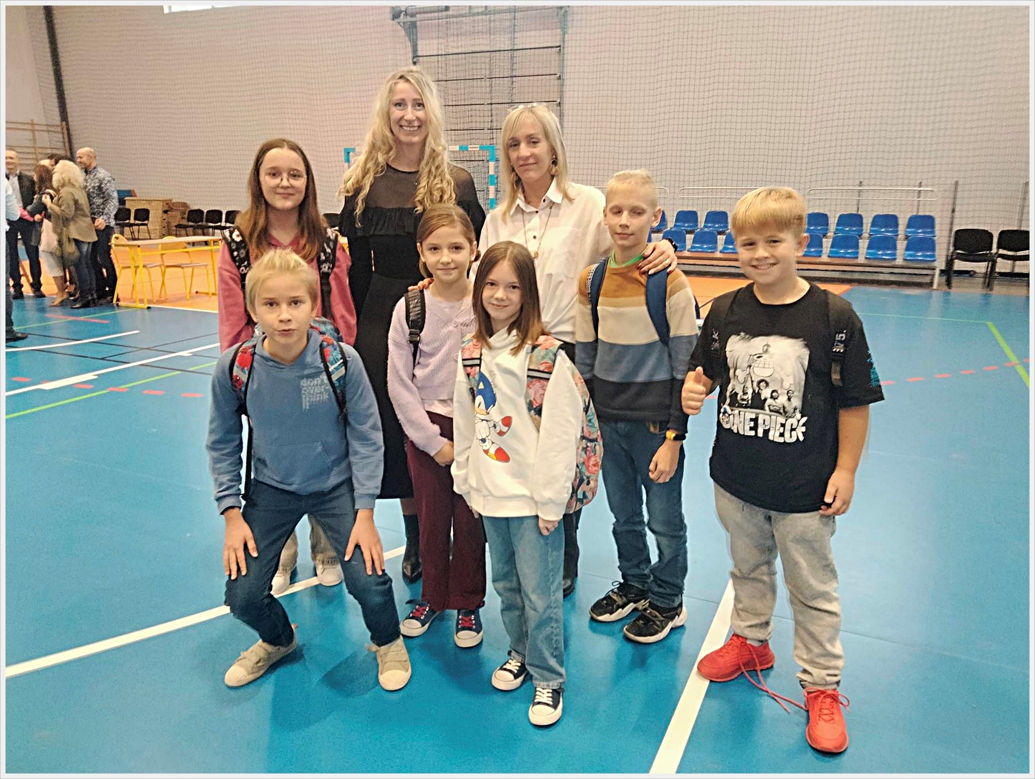 Spotkanie z Pabianickimi olimpijczykami. Grupa siedmiorga uczni&oacute;w i olimpijki Aleksandry Shelton (Socha) wraz z nauczycieleką pozuje do zdjęcia na sali gimnastycznej. Dzieci mają na sobie codzienne ubrania i plecaki, a jedna z dziewczynek nosi bluzę z postacią Sonica. Aleksandra Shelton (Socha) i nauczycielki stoją z tyłu i uśmiechają się. W tle widać niebieskie trybuny, siatkę na ścianie oraz inne osoby rozmawiające przy stolikach.