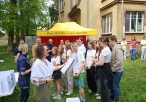 Uczniowie i opiekunowie stoją przy namiocie wolontariatu, słuchają instrukcji do gry miejskiej.