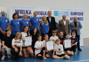 Uczniowie trzymają certyfikaty i stoją obok dorosłych w hali sportowej.