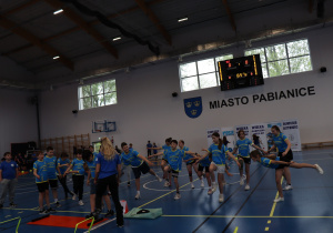Dwóch uczniów biegnie między pachołkami w hali sportowej.