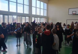 Uczniowie bawią się w halloweenowych przebraniach na sali gimnastycznej; dzieci tańczą i stoją w grupach, w tle widoczni są inni uczniowie siedzący przy ścianie.